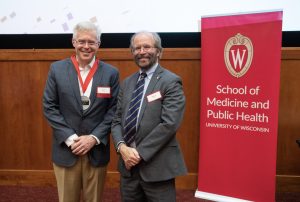 Two men in suits standing in a lecture hall with one wearing a medal recognizing their investiture. 