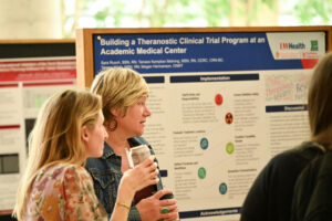 Two women in conversation, with a poster titled "Building a Theranostic Clinical Trial Program at an Academic Medical Center" behind them.